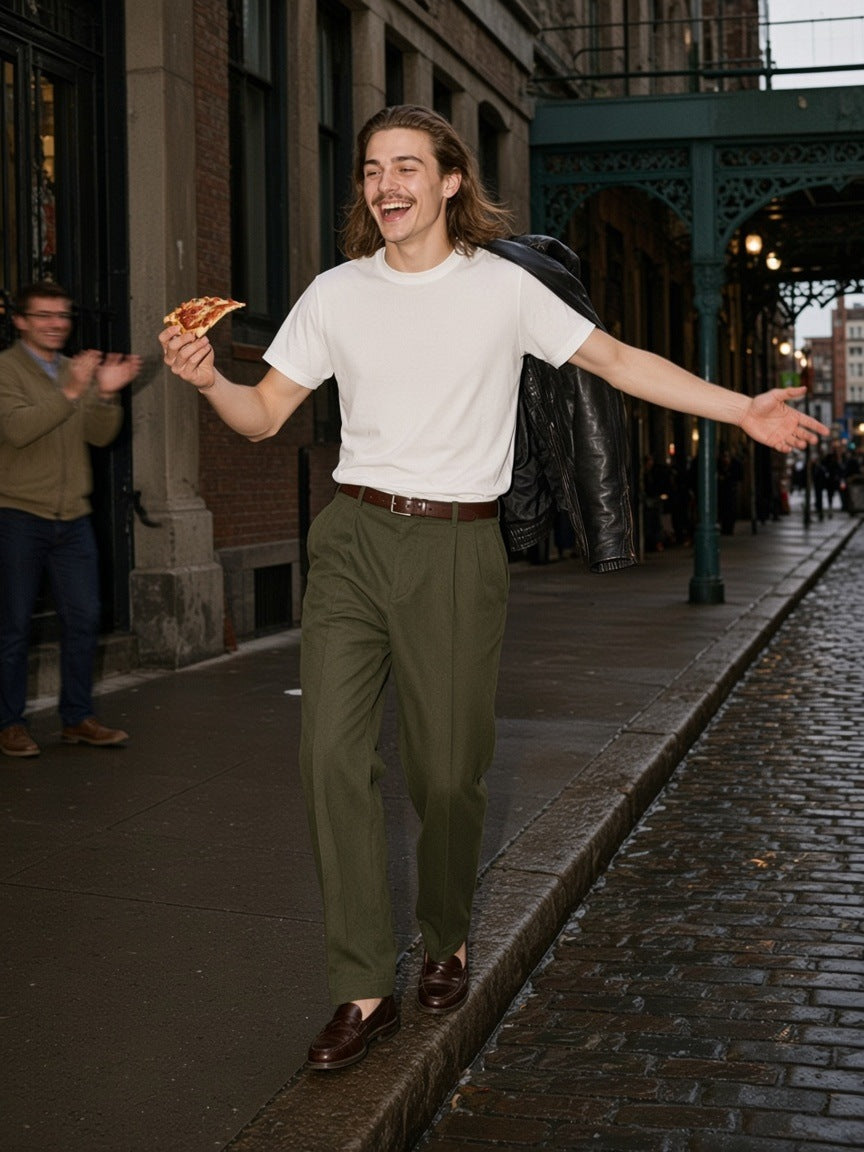 Man in the Marcel Afters Men's Essential Heavyweight Cotton T-Shirt in White with olive trousers, balancing a curb with a pizza slice in lower Manhattan at dusk