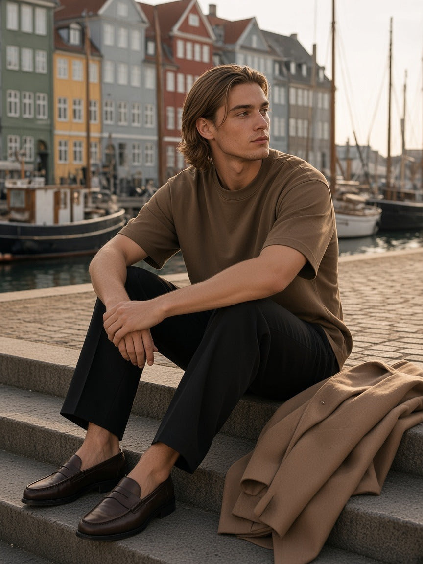 Man in the Heavyweight Drop Shoulder T-Shirt in Coffee with black trousers and loafers, seated on the harbour steps in Copenhagen by day