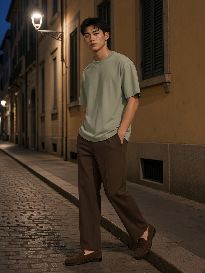 Man in a Grey Green heavyweight drop shoulder t-shirt and brown trousers on a lamplit cobbled street at dusk