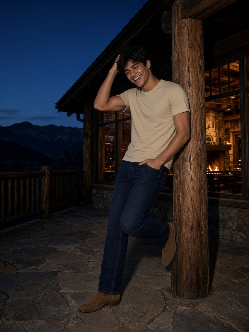 Man in a Sand Color heavyweight earth tone t-shirt and dark jeans laughing against a chalet terrace post at dusk