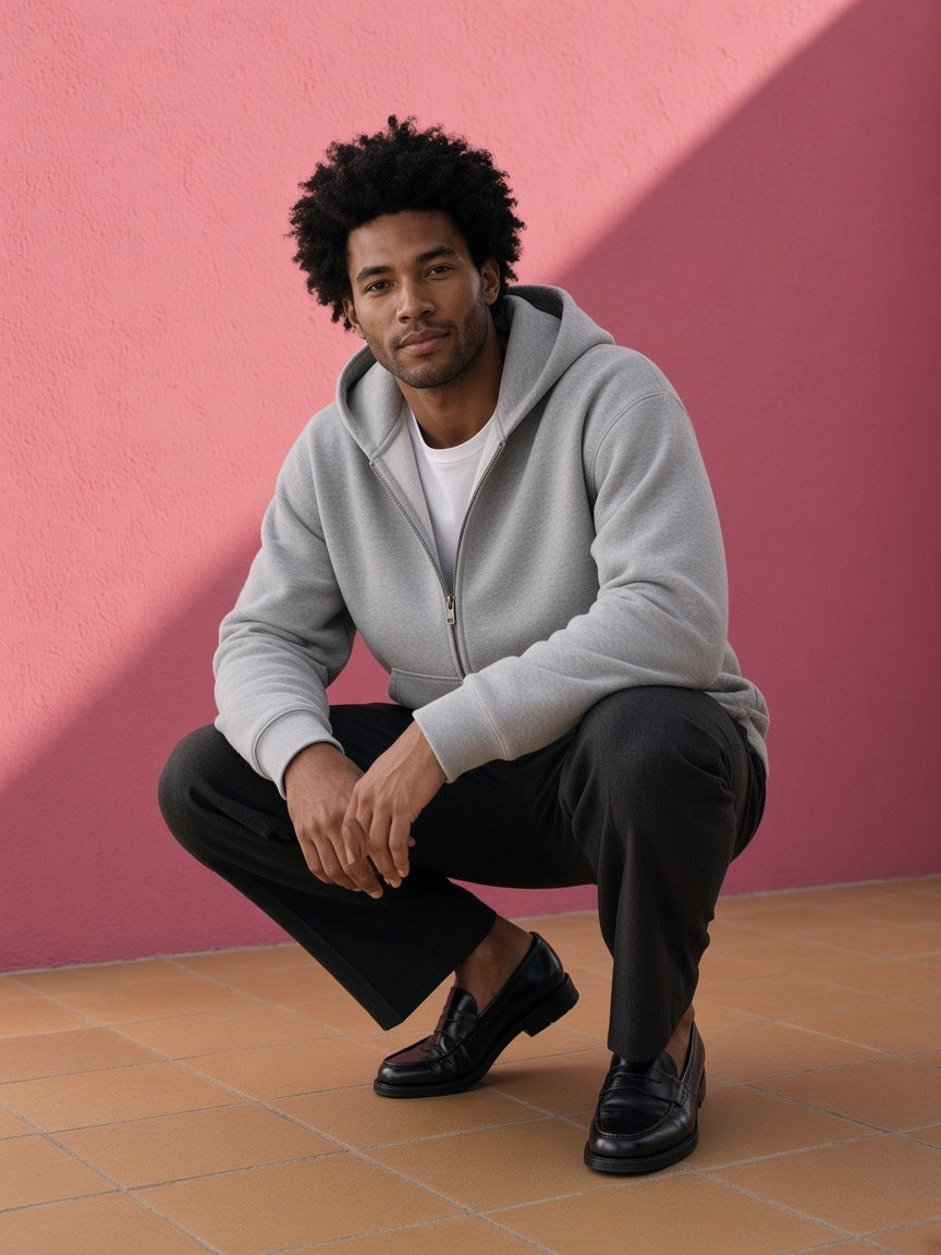 Man in the Men's Heavyweight Full Zip Boxy Hoodie in Flower Gray over a white tee with black trousers and loafers, crouched by a pink plaster wall in Mexico City by day