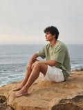 Man in the Men's Heavyweight Vintage Washed Frayed T-Shirt in Gray Green with white shorts, seated on sea rocks at dawn