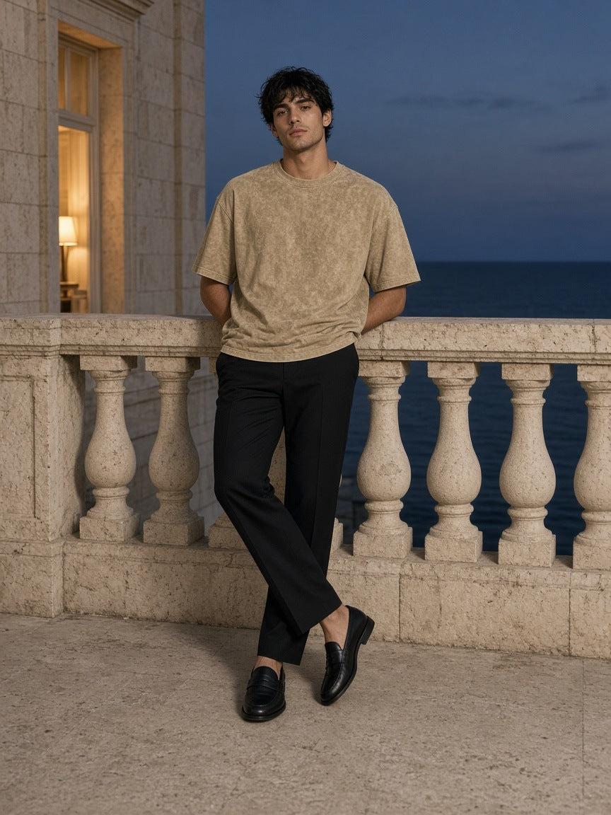 Man in the Men's Snow Washed Heavyweight Oversize T-Shirt in Khaki leaning on a limestone hotel terrace above the sea at night