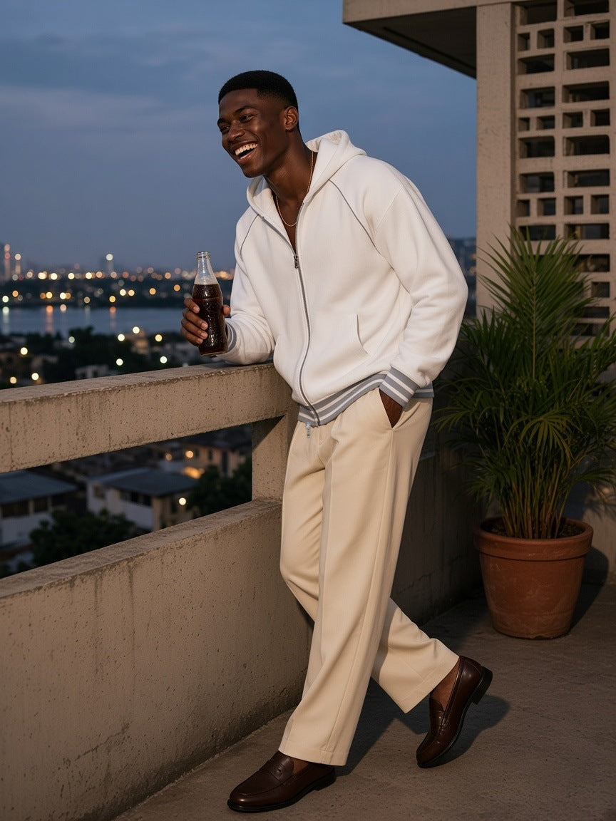 Man in the Men's Striped Trim Zip-Up Hoodie in White with cream trousers and loafers, laughing at a concrete balcony rail over the lagoon in Lagos at dusk
