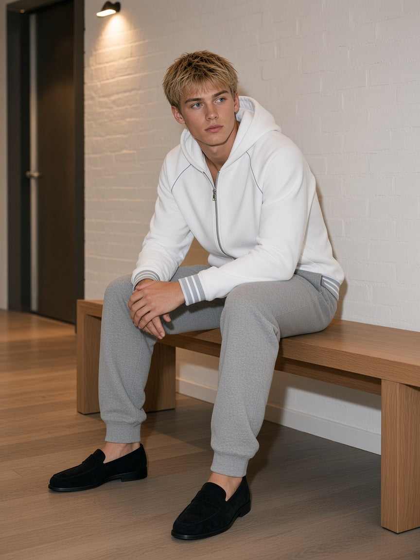 Man in the Men's Striped Trim Zip-Up Hoodie in White with grey joggers, seated on an oak bench against white brick in Stockholm