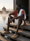 Man in the Mesh-Lined Quick-Dry Performance Shorts in Grayish White with a rust camp shirt and suede clogs, towel on shoulder, seated on the Aarhus harbour sauna jetty at dawn