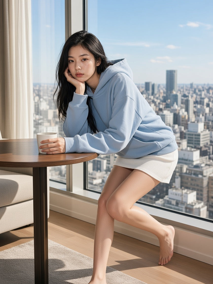 Woman in the Mineral Wash Boxy Hoodie in Haze Blue with a white knit skirt, barefoot in a hotel suite over Tokyo in the morning