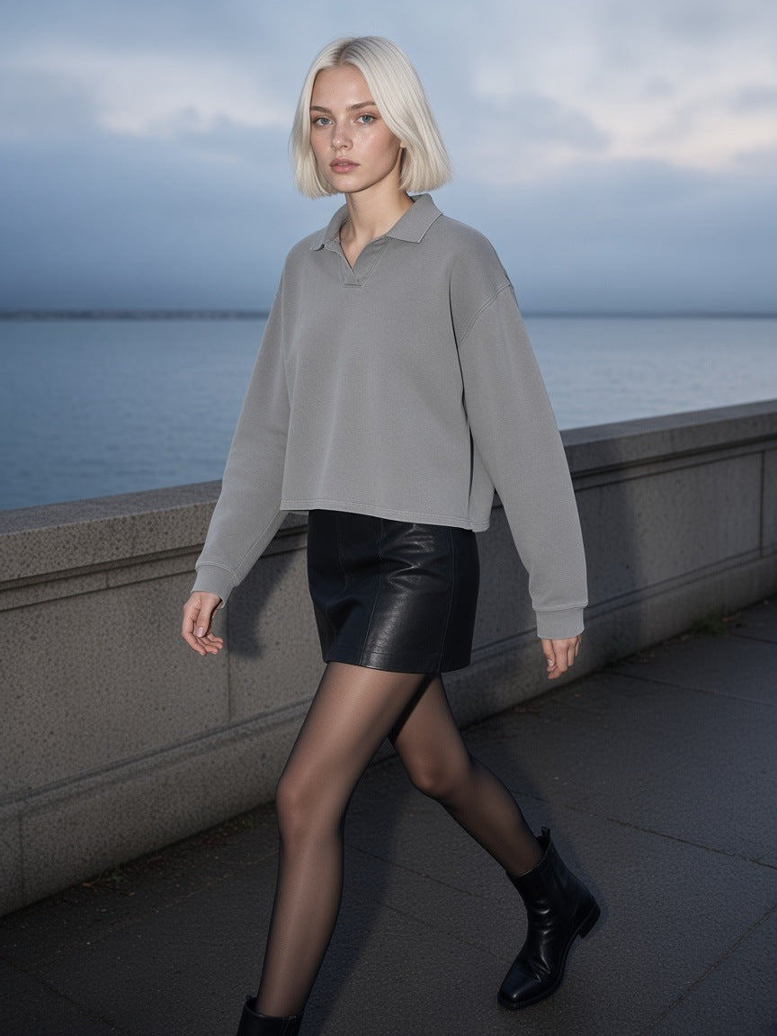 Woman in the Marcel Afters Mineral Wash Pullover Sweatshirt in Medium Gray with a black leather mini, sheer tights and ankle boots, walking a seawall promenade
