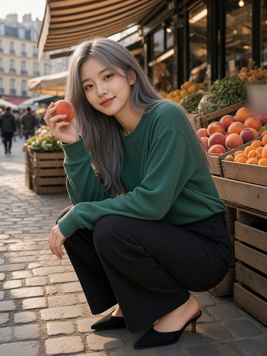 Woman in the Mineral Wash Waffle Oversized T-Shirt in Blackish Green with wide black trousers and kitten-heel mules, crouched by the peach crates at Marché d'Aligre, Paris in the morning