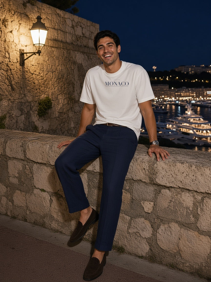 Man in the Monaco Heavyweight Drop Shoulder T-Shirt in White with the navy Monaco city plate, on a yacht deck in the harbour of Monaco at night
