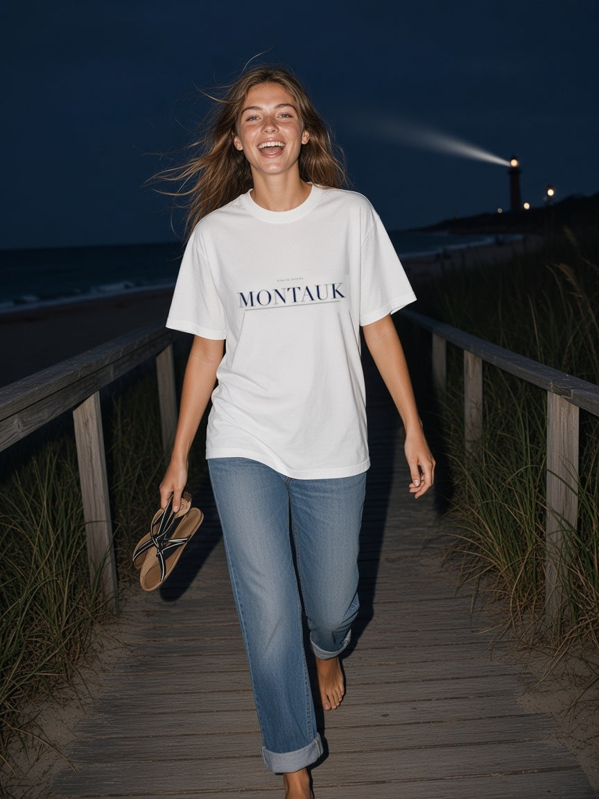 Woman in the Montauk Heavyweight Drop Shoulder T-Shirt in White with the navy Montauk city plate, on a boardwalk below the lighthouse in Montauk at night
