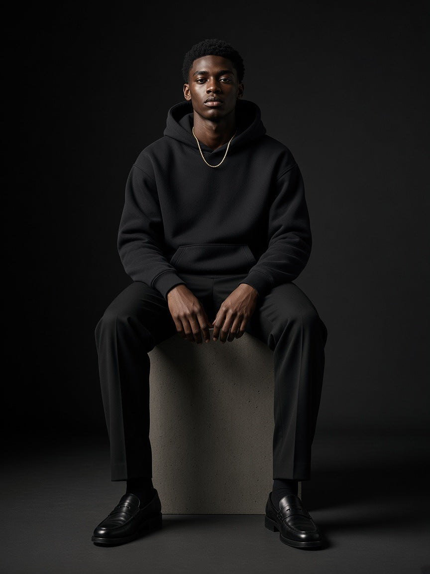 Man in the Marcel Afters Oversize Heavyweight Fleece Hoodie in Black Beauty, seated on a concrete plinth in a charcoal studio