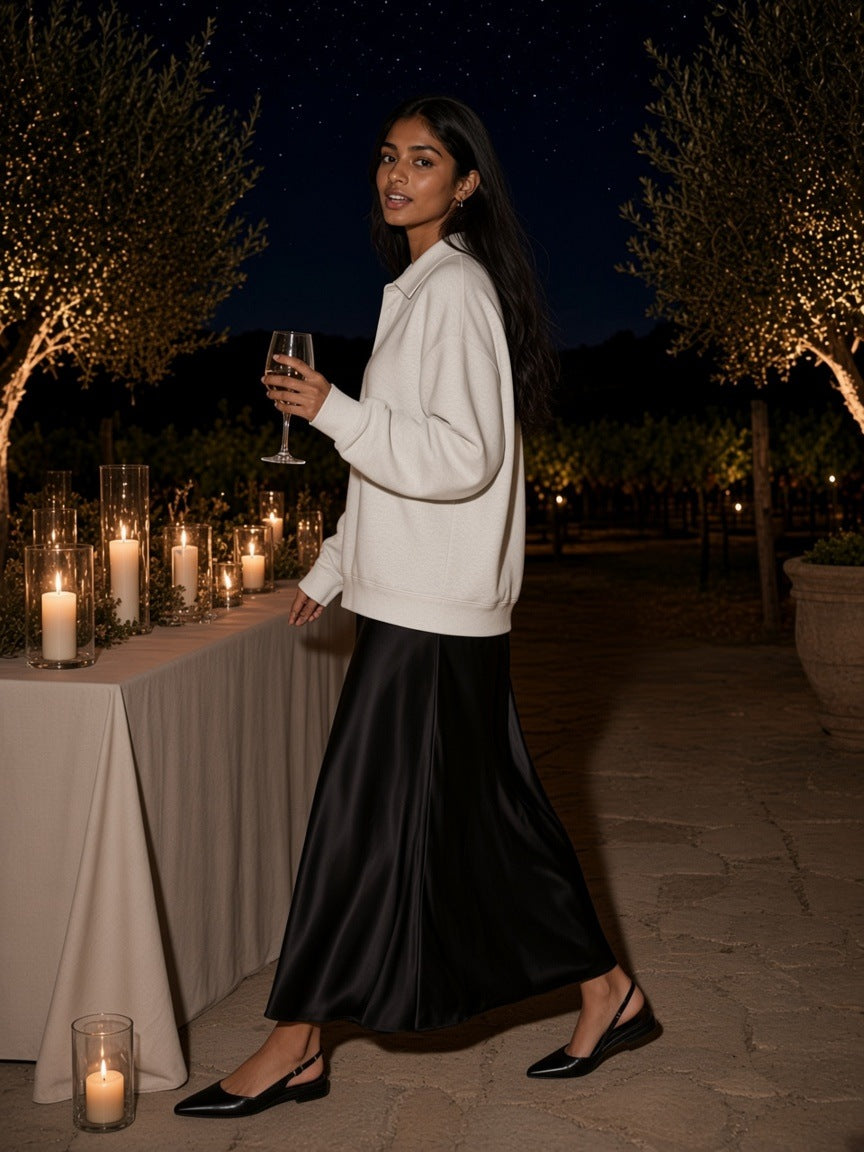 Woman in the Oversized Collared Sweatshirt in Oat Gray with a black satin skirt, raising a glass on a candlelit vineyard terrace at night