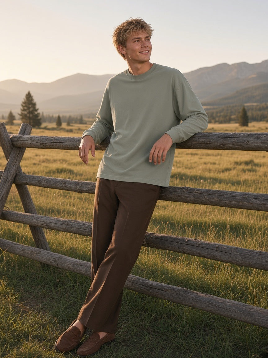 Man in the Oversized Long Sleeve T-Shirt in Gray Green with chocolate trousers and suede loafers, leaning on a ranch fence in Montana in the morning