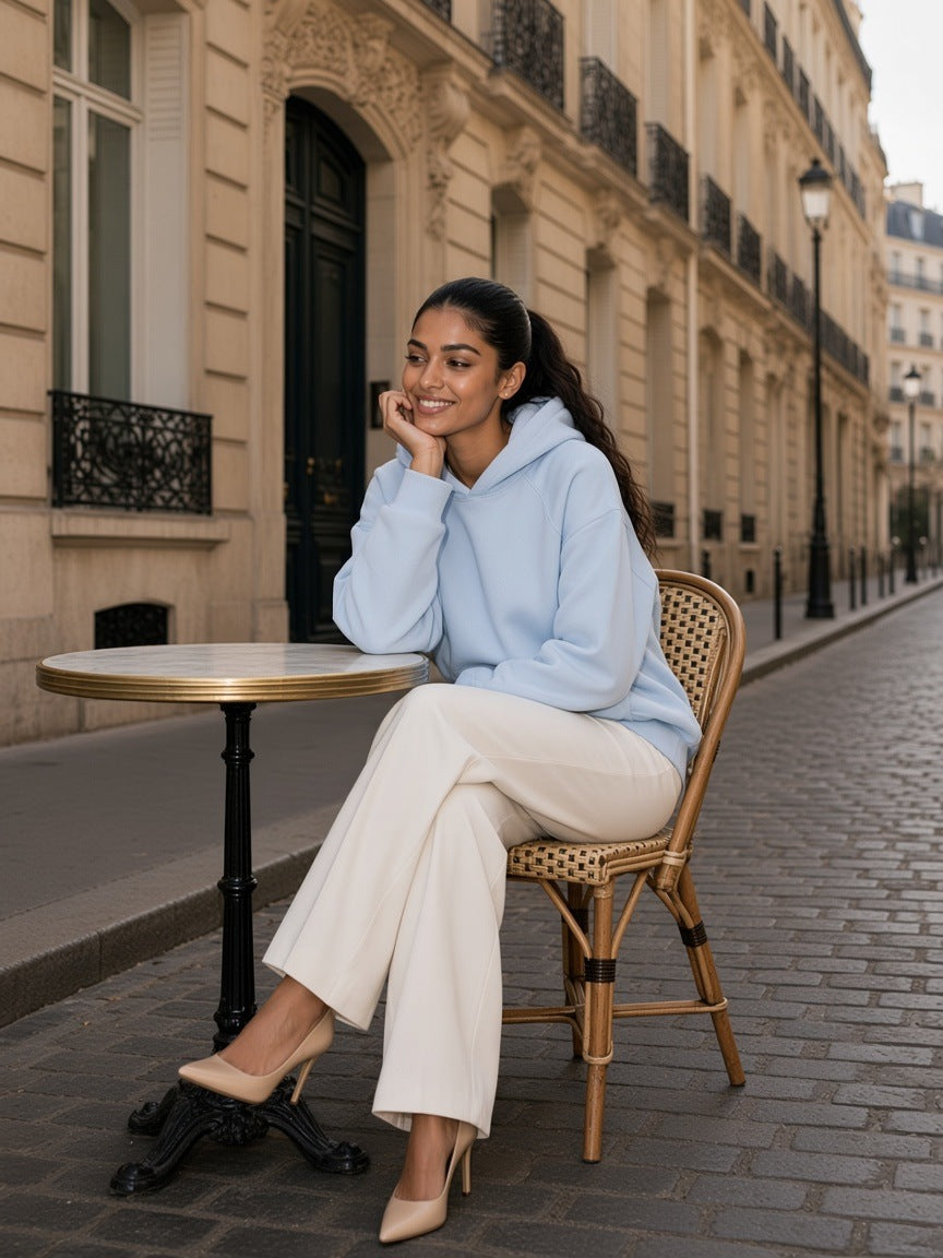 Woman in the Oversized Raglan Pullover Hoodie in Light Blue with white trousers and heels, walking a Haussmann street in Paris by day