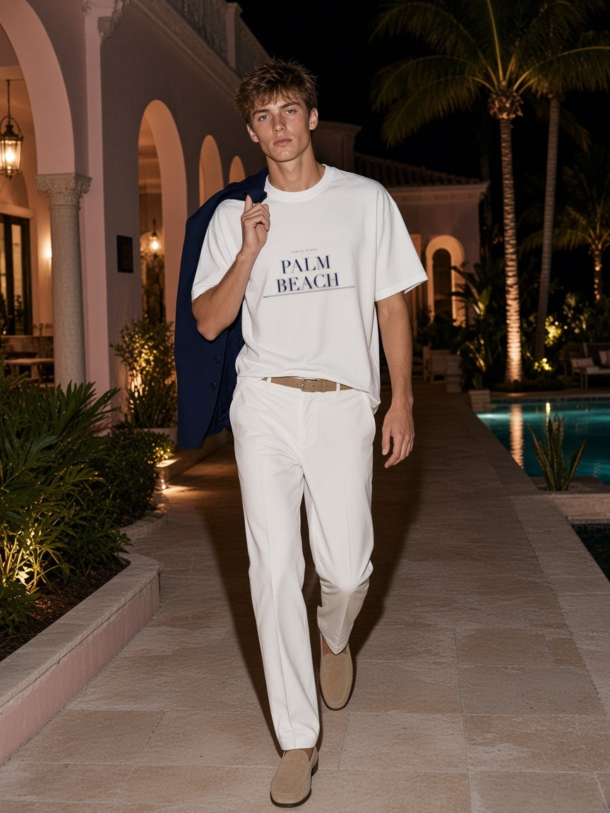 Man in the Palm Beach Heavyweight Drop Shoulder T-Shirt in White with the navy Palm Beach city plate, walking past a pink oceanfront mansion in Palm Beach at night