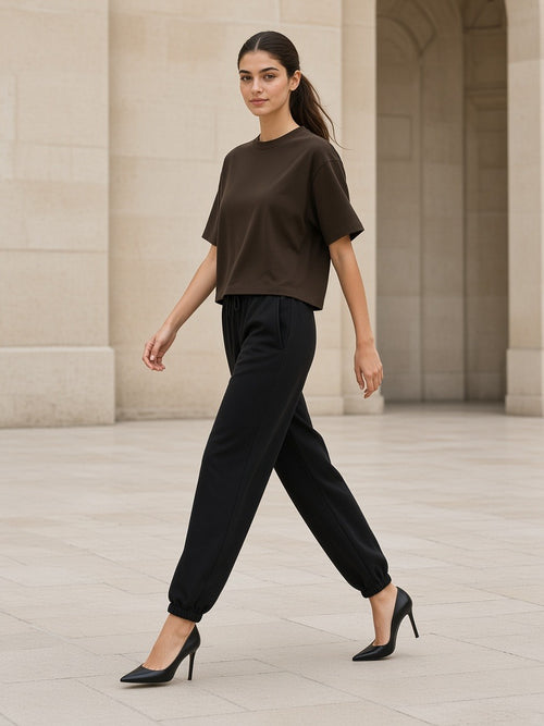 Woman in the Heavyweight Boxy T-Shirt in Coffee with black loose tapered joggers striding across a limestone Paris courtyard in heels