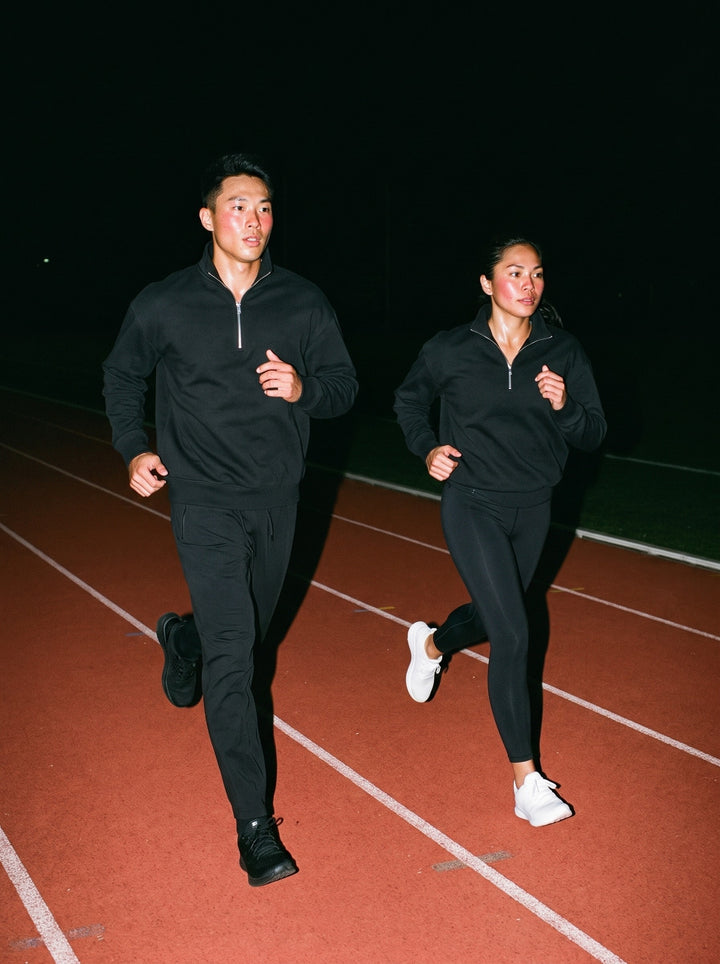 Two people in the Quarter-Zip Drop Shoulder Sweatshirt, a man in Black and a woman in Black, running strides on a track at night