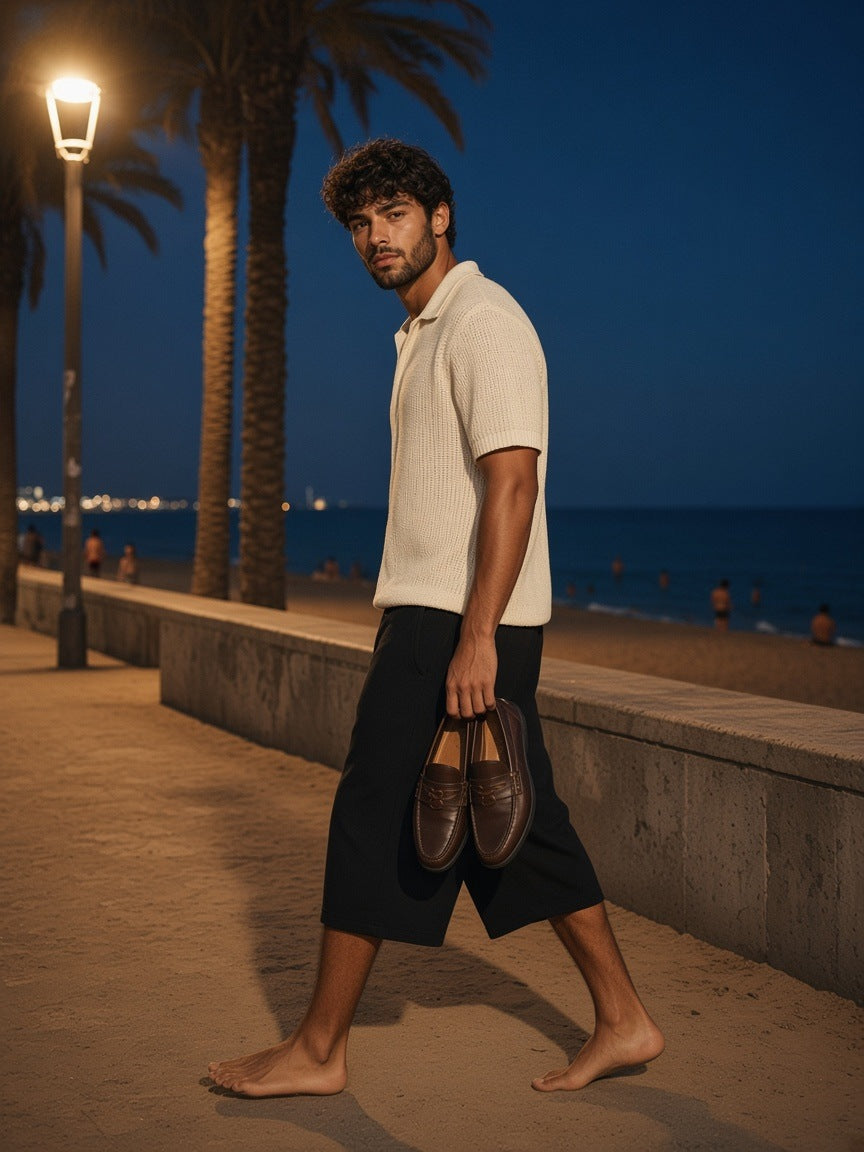 Man in the Relaxed Drawstring Three-quarter Sweatpants in Black with a cream knit polo, carrying his loafers along the Barcelona boardwalk at night