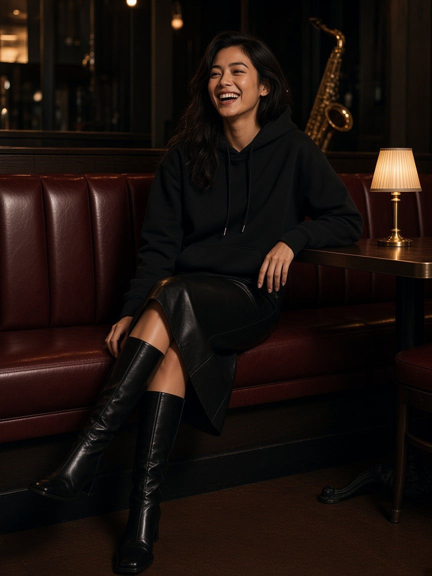 Woman in the Relaxed Pullover Hoodie in Black with a black leather midi skirt and knee boots, laughing in a jazz club booth at night