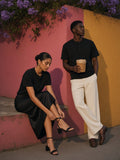 Two people in the Short Sleeve Henley Shirt in Black, one tying a sandal on a ledge and one leaning on a pink and ochre wall in Mexico City at dusk