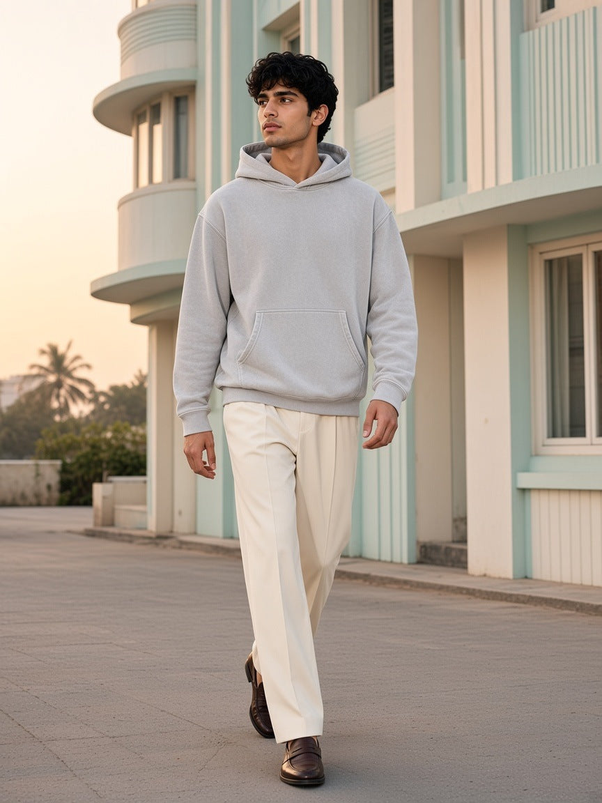 Man in the Marcel Afters Snow Wash Fleece Oversize Hoodie in Light Gray with cream trousers and loafers, past an Art Deco hotel in Mumbai at dawn
