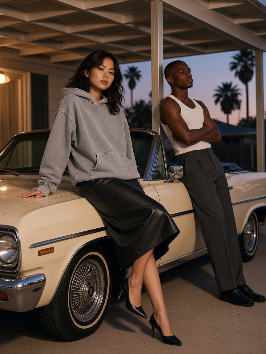 Two people at a Palm Springs carport at dusk, one wearing the Snow Wash Hand-Frayed Heavyweight Hoodie in Light Gray with a black leather midi, perched on a vintage car