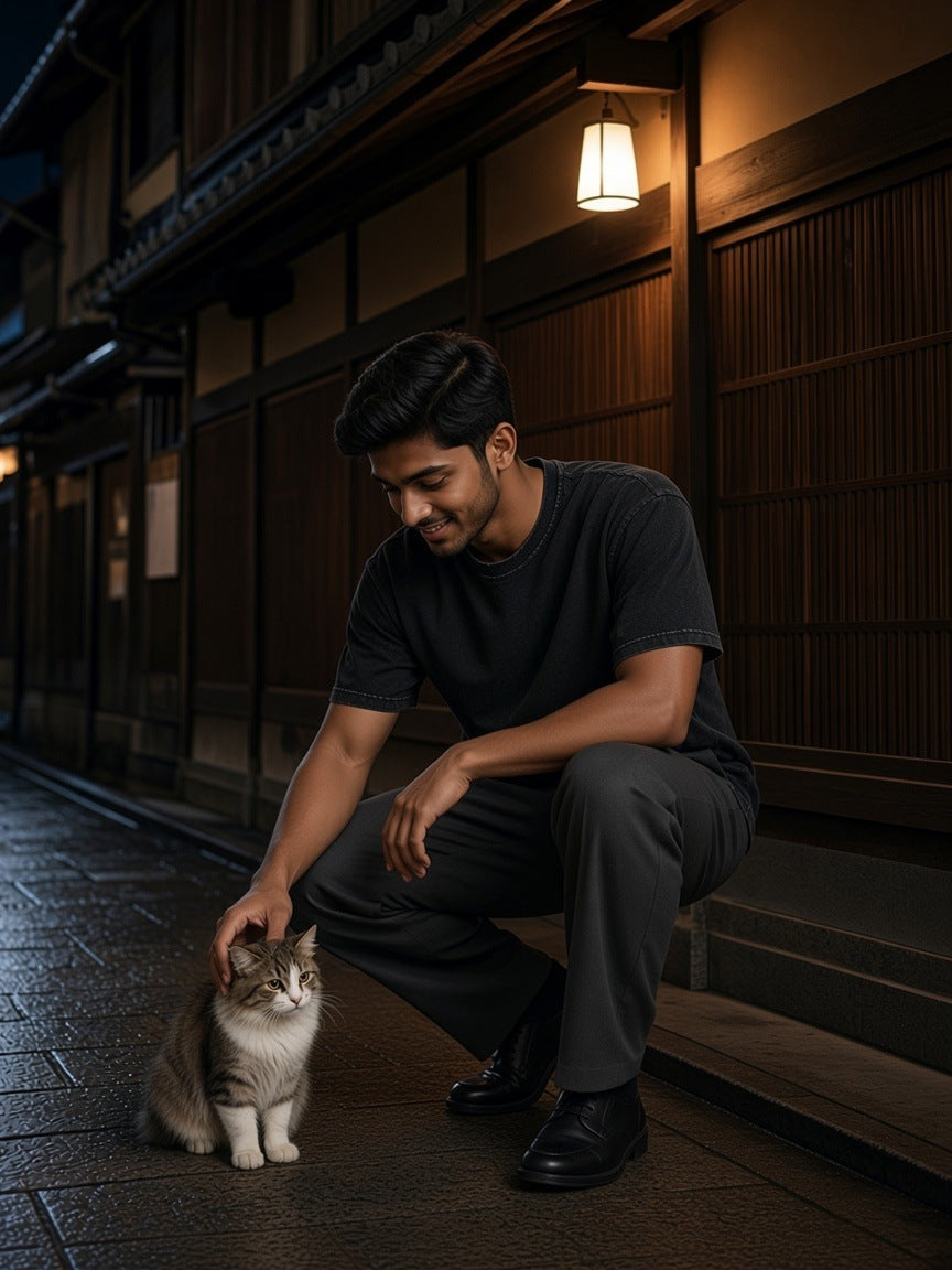 Man in the Snow Washed Raw-Edge Neckline T-Shirt in Black crouching to pet a cat in a lantern-lit timber lane