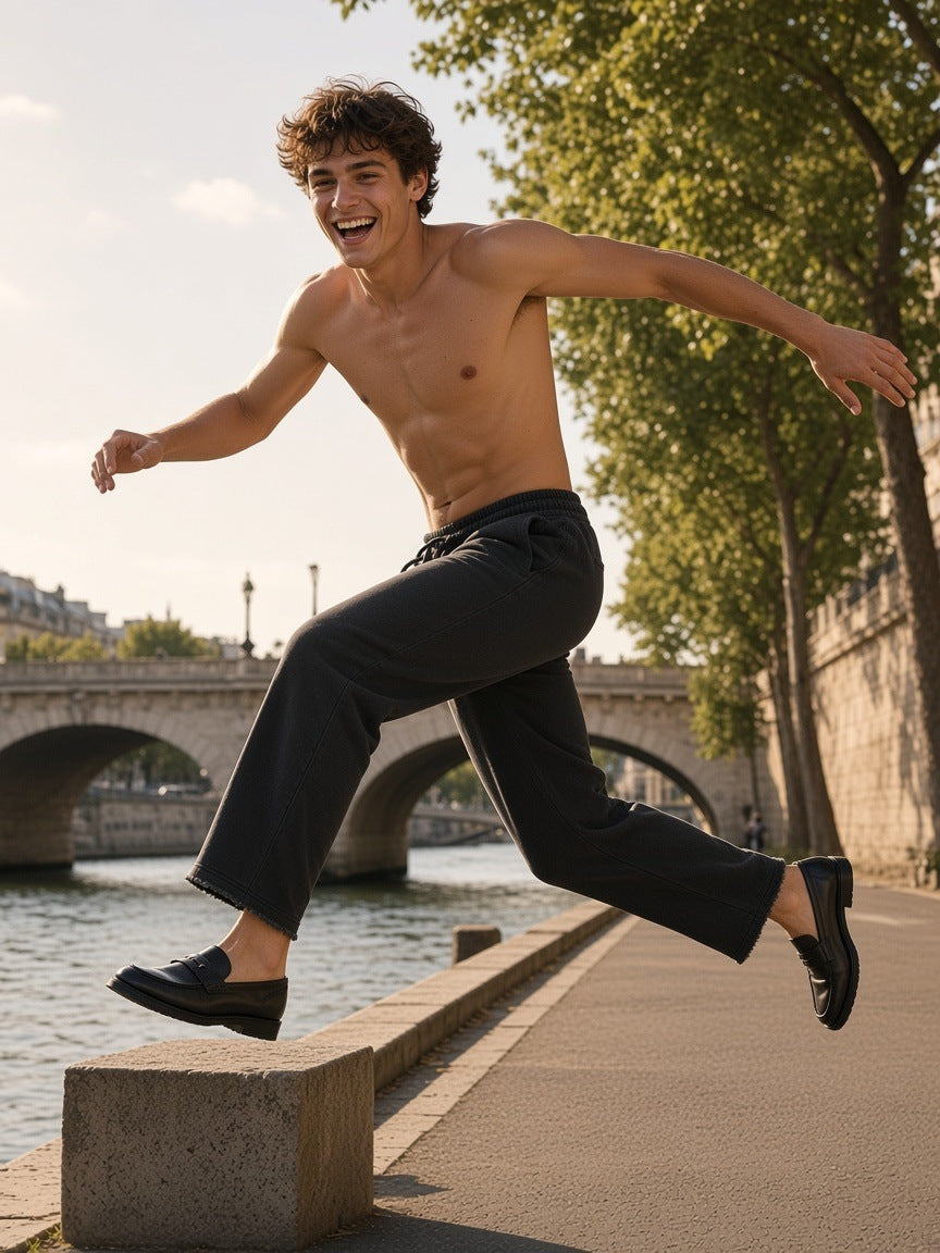 Man in the Snow Washed Raw Edge Sweatpants in Black, shirtless, leaping between stone blocks on a Seine quay at golden hour