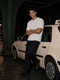 Man in a white Sorona quick-dry t-shirt and black trousers leaning on a cream taxi under an iron bridge at night