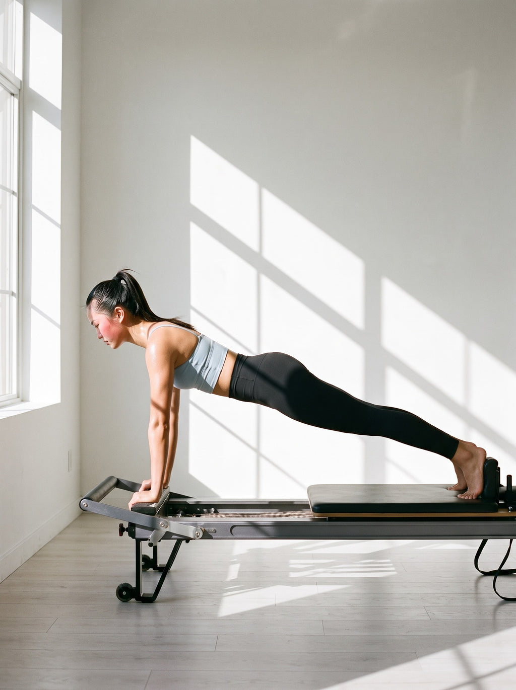 Woman in the Spaghetti Strap Yoga Sports Bra in Haze Blue, holding a plank on a pilates reformer in a sunlit studio