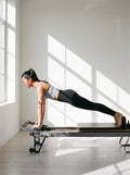 Woman in the Spaghetti Strap Yoga Sports Bra in Haze Blue, holding a plank on a pilates reformer in a sunlit studio