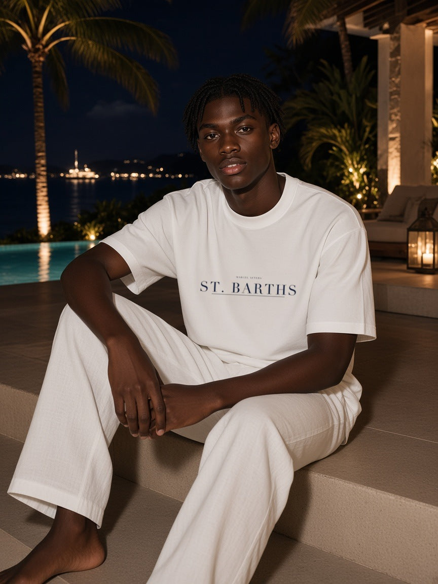 Man in the St. Barths Heavyweight Drop Shoulder T-Shirt in White with the navy St. Barths city plate, seated on villa pool steps in St. Barths at night