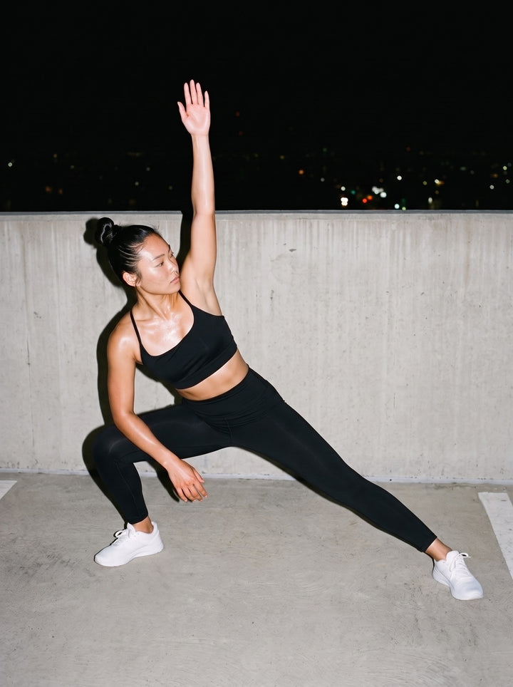 Woman in the Strappy Criss Cross Yoga Sports Bra in Black, in a side lunge stretch on a rooftop at night