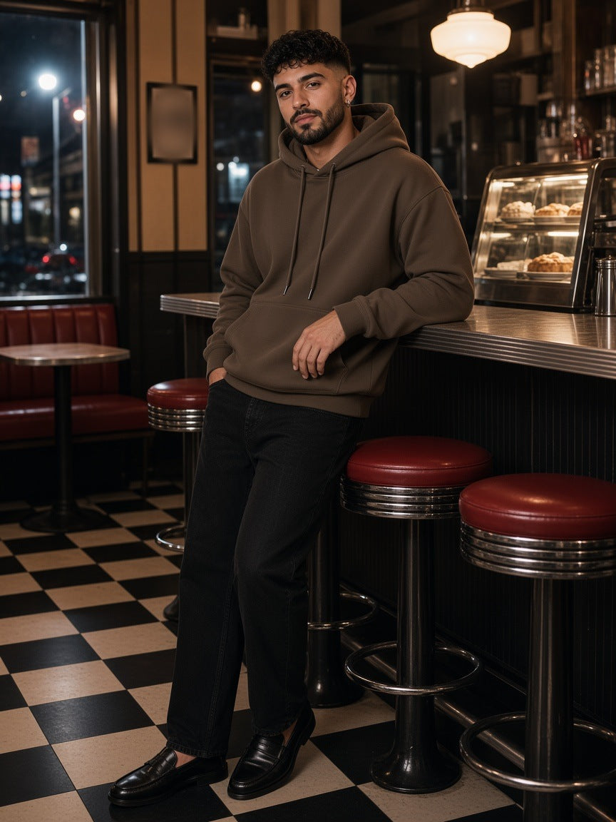 Man in the Streetwear Fleece Hoodie in Dark Brown with black jeans and penny loafers, leaning on the counter of a Bowery diner in New York at night