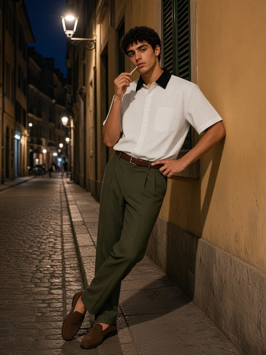 Man in the Striped Boxy Contrast Collar Shirt in White, tucked into olive pleated trousers, leaning on an ochre wall in Milan at night