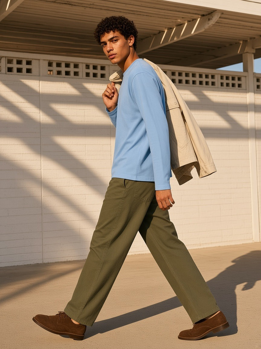 Man in the Sunfade Boxy Long Sleeve T-Shirt in Sky Blue with olive twill trousers and brown suede derbies, a jacket over one shoulder, walking through a Palm Springs carport's shadow grid at golden hour
