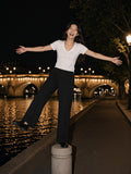 Marcel Afters Tencel Slim Fit V-Neck T-Shirt in White, worn by a woman, arms wide on a stone bollard beside a lit night river in a white v-neck and black trousers