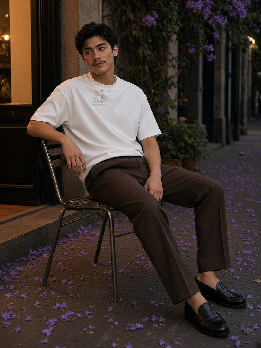 Man in The Dogs Made The Album Heavyweight Drop Shoulder T-Shirt in White seated on a café chair on a jacaranda street in Mexico City at night
