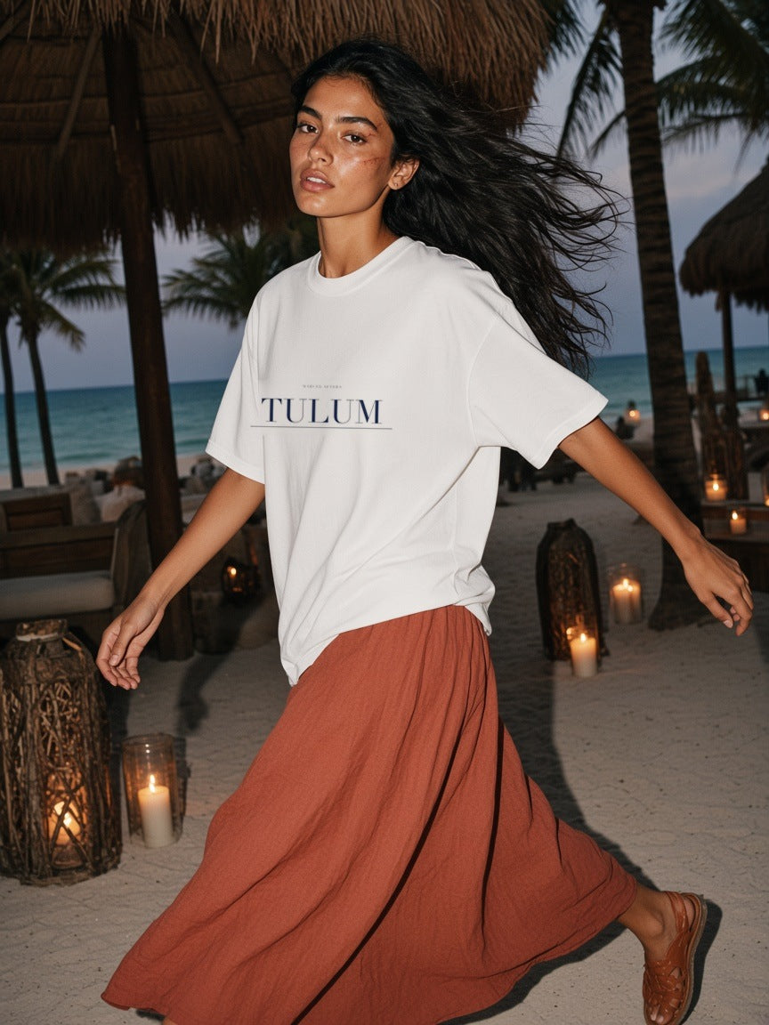 Woman in the Tulum Heavyweight Drop Shoulder T-Shirt in White with the navy Tulum city plate, on a candlelit beach path under the palms in Tulum at night