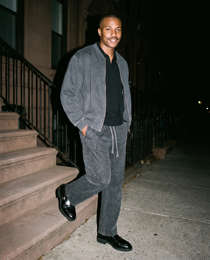 Man in Black heavyweight oversized polo shirt under a gray suede jacket, with gray drawstring trousers, on a stoop at night