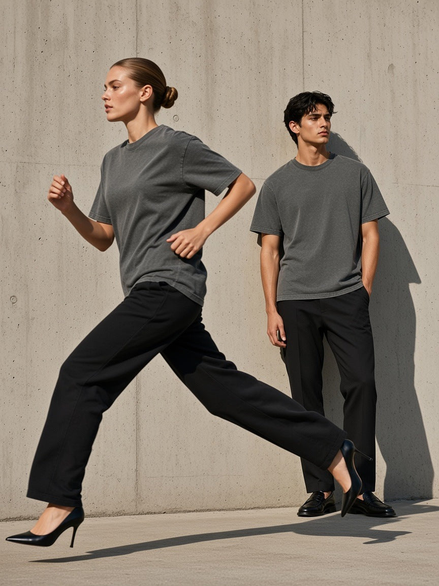 Two people in the Marcel Afters Mineral Wash Oversized T-Shirt in Charcoal Gray, a woman running in heels past a raw concrete wall, a man standing behind
