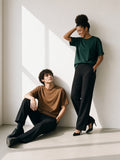 Two people in the Oversized Drop Shoulder T-Shirt in Dark Green and Brown, a woman laughing and a man sitting against a sunlit plaster wall in the morning