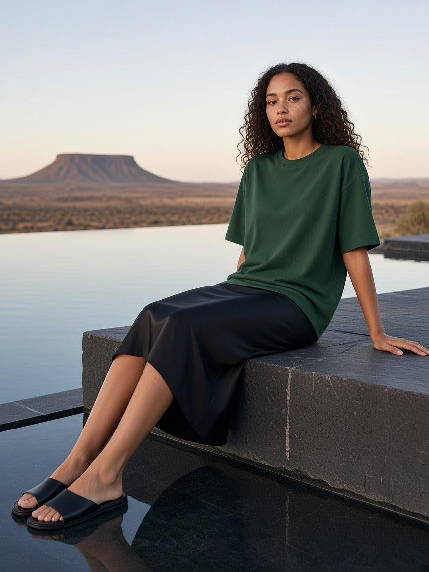Woman in the Marcel Afters Oversized Drop Shoulder T-Shirt in Dark Green, seated on the edge of a dark stone infinity pool in the desert