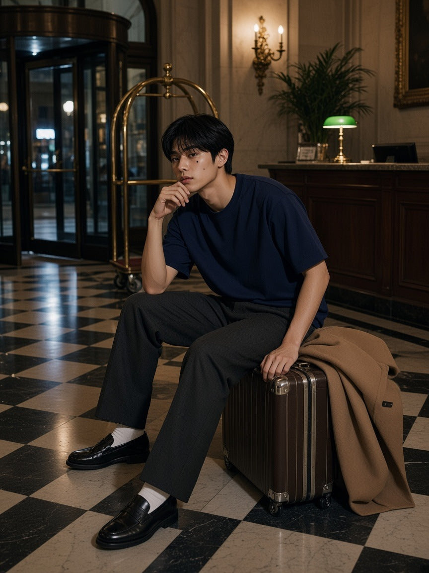 Man in the Oversized T-Shirt in Navy Blue seated on a suitcase in a grand hotel lobby late at night