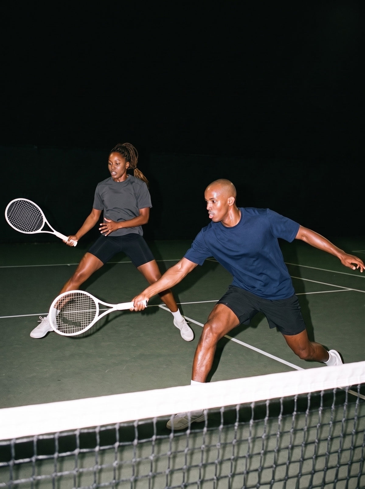Two people in the Sorona Quick-Dry Loose Fit Boxy T-Shirt, a man in Navy Blue and a woman in Dark Gray, mid-rally on a tennis court at night