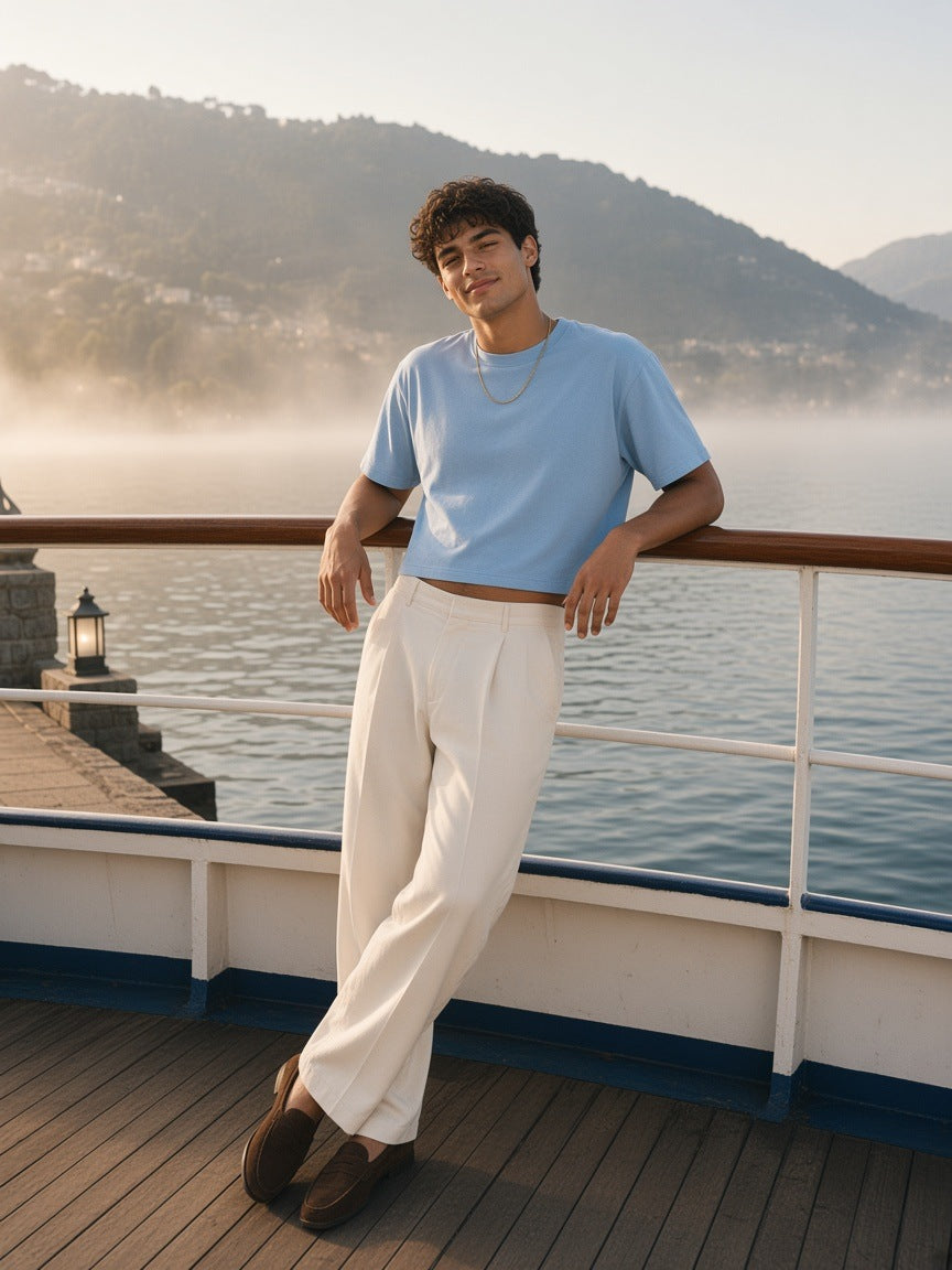 Man in the Vintage Wash Sunfade Boxy T-Shirt in Sky Blue with cream pleated trousers and suede loafers, leaning on a Lake Como ferry rail in the morning
