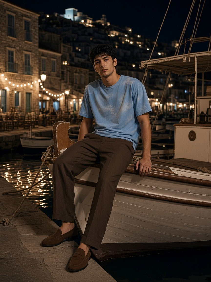 Man in the Vintage Wash Sunfade Boxy T-Shirt in Sky Blue on the bow of a wooden boat in Hydra harbour at night