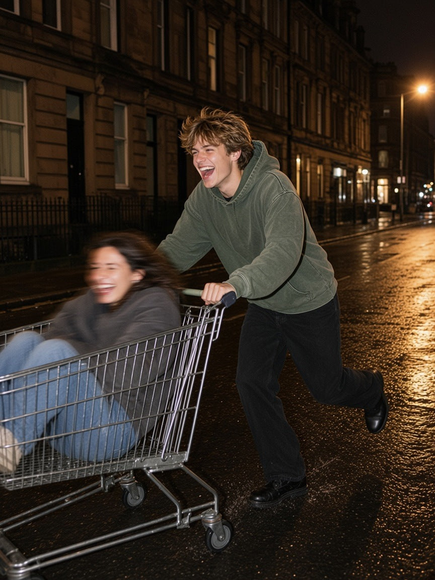 Two people in the Vintage Washed Frayed-Hem Hoodie: a man in Gray Green racing a trolley with a woman riding in it down a wet Glasgow street at 4am