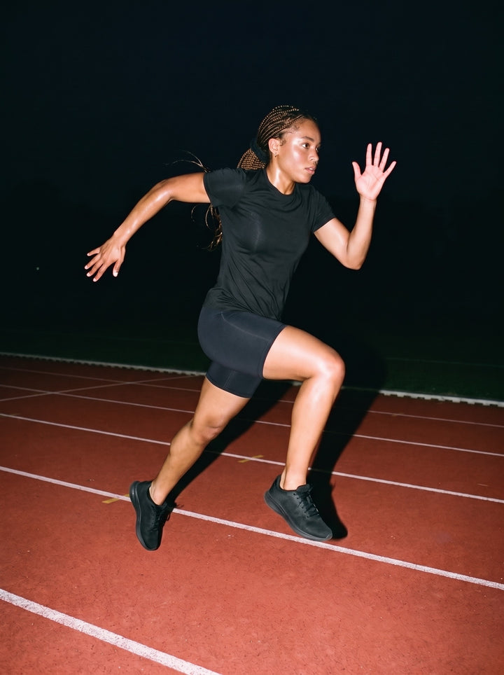 Woman in the Women's Slim Fit Performance T-Shirt in Black, sprinting down a running track at night